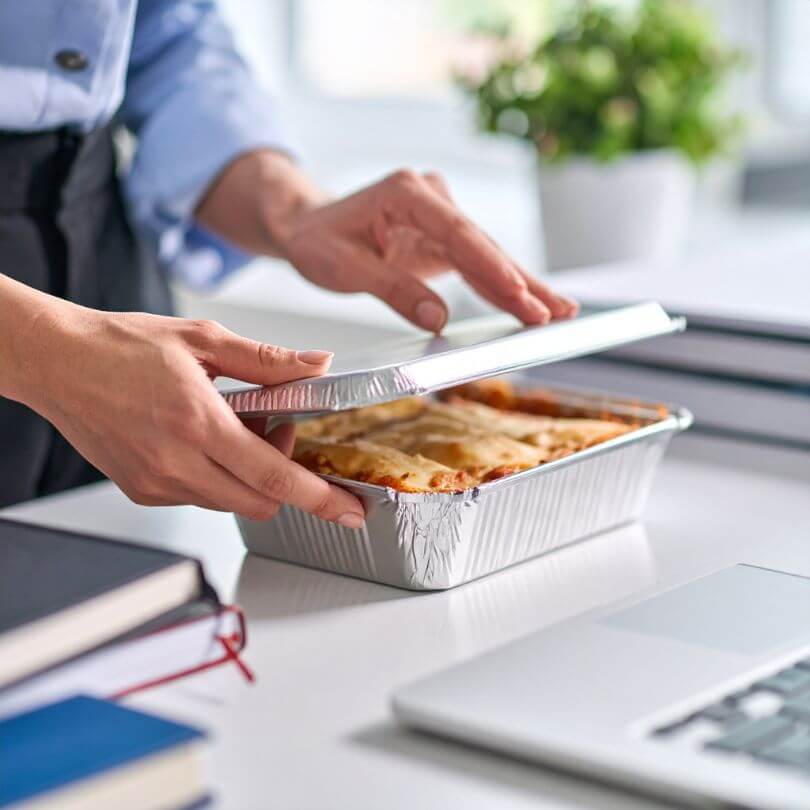 Donna che apre un contenitore di lasagna proteica vegetariana pronta su un tavolo di lavoro.
