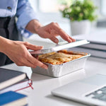 Donna che apre un contenitore di lasagna proteica vegetariana pronta su un tavolo di lavoro.