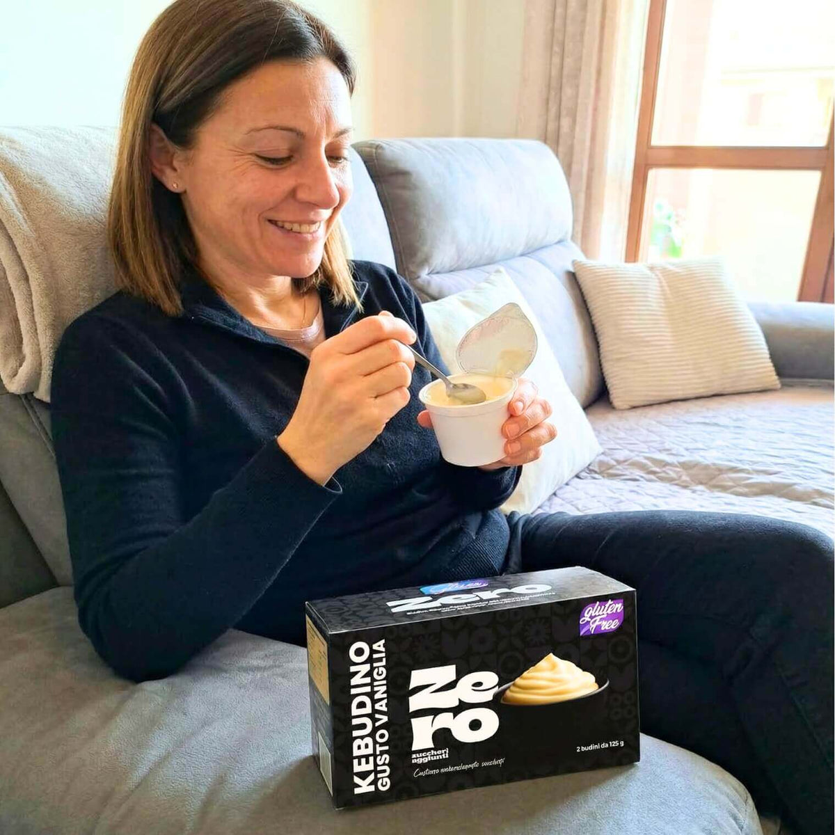 Donna che gusta Kebudino proteico alla Vaniglia su divano, con budino e confezione in vista.